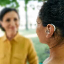 Adult woman with a hearing impairment uses a hearing aid to communicate with her female friend at city park. Hearing solutions